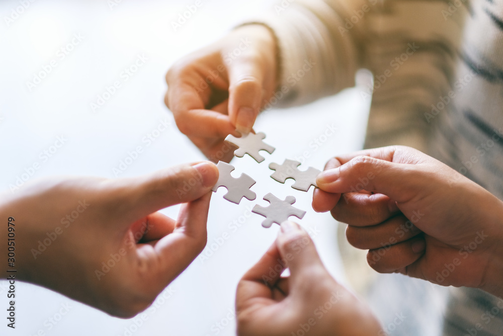 Closeup image of many people holding and putting a piece of white jigsaw puzzle together - obrazy, fototapety, plakaty 