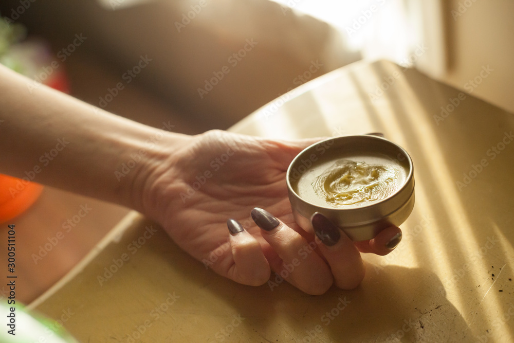 Woman holds a metal tin of homemade all natural organic homeopathic CBD balm from small business for pain and anxiety management and is backlit by window light - obrazy, fototapety, plakaty 