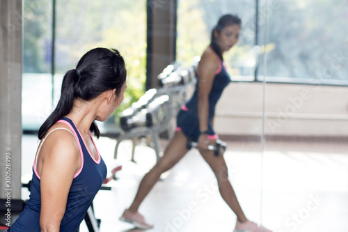 Young woman practicing with dumbbells in front of mirror in gym. Girl strengthens in fitness club.
