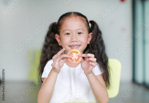 Smiling little Asian child girl holding a piece of sliced tomato. Kid eating healthy food concept. Focus at tomato in children hands.