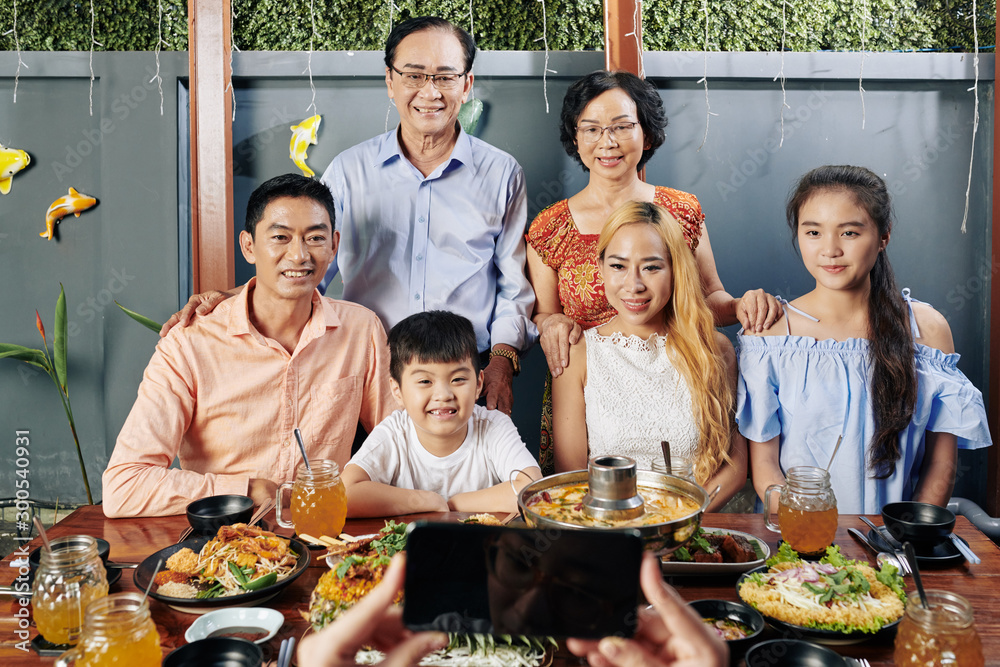 Happy big Vietnamese family posing at big dinner table for photo on smartphone - obrazy, fototapety, plakaty 