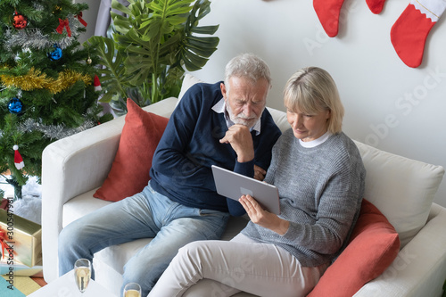 senior elderly caucasian old man and woman sitting on sofa, using and play internet tablet together in living room that decorated for christmas festival day in the morning, retirement family concept