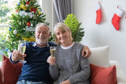 senior elderly caucasian old man and woman holding glass of champagne celebrate together in living room that decorated with christmas tree for christmas festival day, retirement lifestyle concept