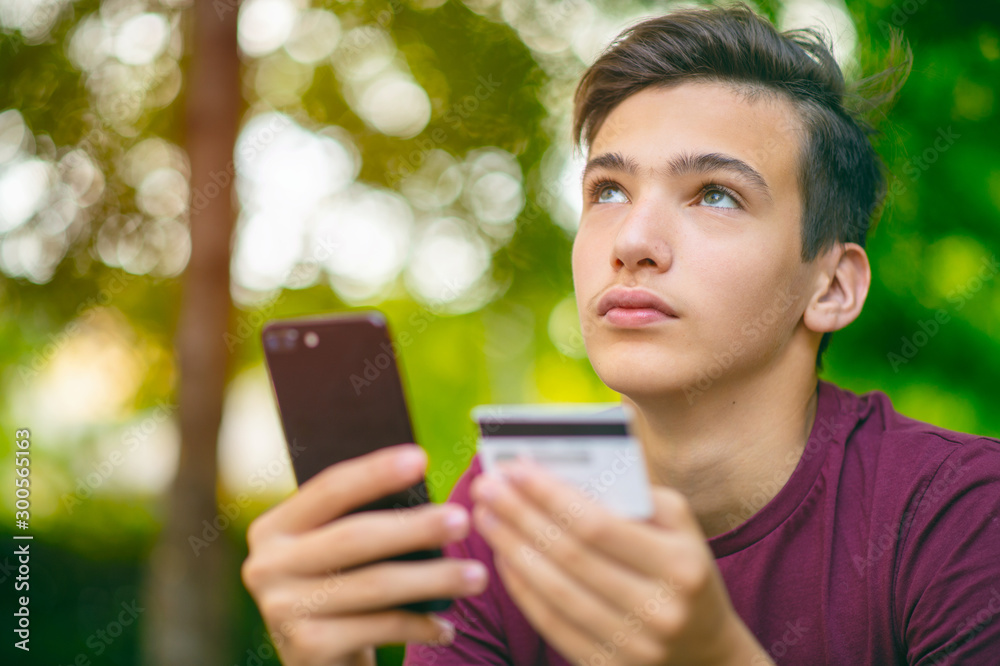 Teenage boy with a credit card and phone, outdoors. Unhappy young man is using smart phone and bank card . Handsome smiling white guy holds bank  card and cell phone