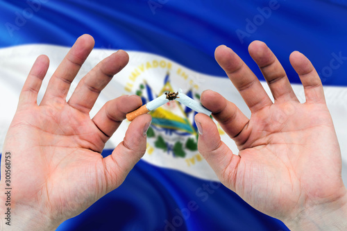 El Salvador quit smoking cigarettes concept. Adult man hands breaking cigarette. National health theme and country flag background.