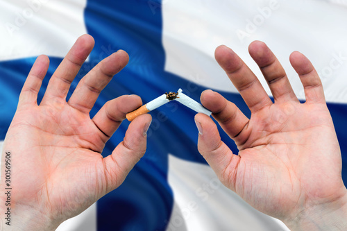Finland quit smoking cigarettes concept. Adult man hands breaking cigarette. National health theme and country flag background.