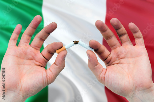 Italy quit smoking cigarettes concept. Adult man hands breaking cigarette. National health theme and country flag background.