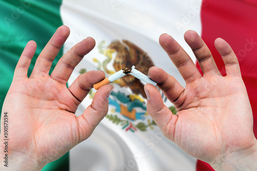 Mexico quit smoking cigarettes concept. Adult man hands breaking cigarette. National health theme and country flag background.