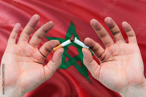 Morocco quit smoking cigarettes concept. Adult man hands breaking cigarette. National health theme and country flag background.