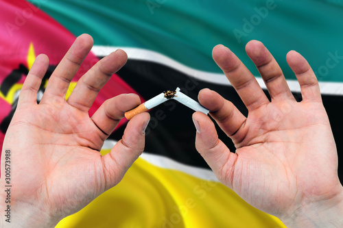 Mozambique quit smoking cigarettes concept. Adult man hands breaking cigarette. National health theme and country flag background.