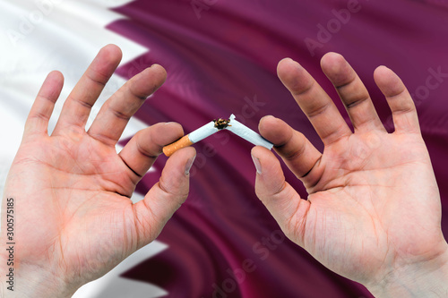 Qatar quit smoking cigarettes concept. Adult man hands breaking cigarette. National health theme and country flag background.