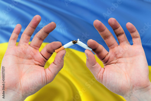 Ukraine quit smoking cigarettes concept. Adult man hands breaking cigarette. National health theme and country flag background.