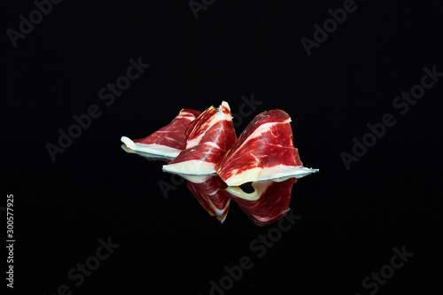 studio photo of a series of slices of 100% Iberian acorn-fed ham folded on black background with reflection
