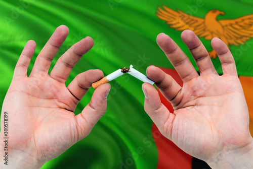 Zambia quit smoking cigarettes concept. Adult man hands breaking cigarette. National health theme and country flag background.