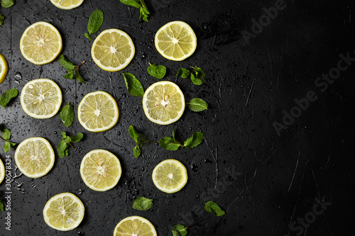 Lemon slices and green mint leaves on black background with water drops, top view. Fresh tropical fruit, yellow citrus, flat lay, copy space