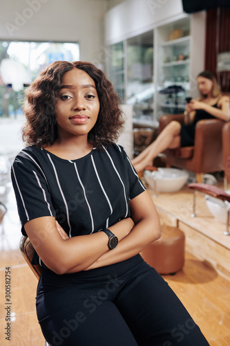 Pretty young Black woman sitting with arms folded and waiting for beautician in beauty salon