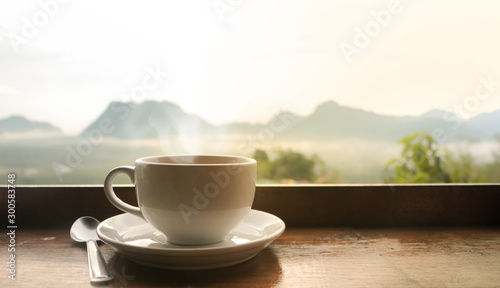 White ceramic coffee cup on wooden table in morning with sunlight over blurred mountains landscape 