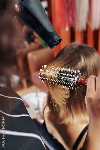 Professional hairdresser using round brush and dryer when drying hair of female client in beauty salon