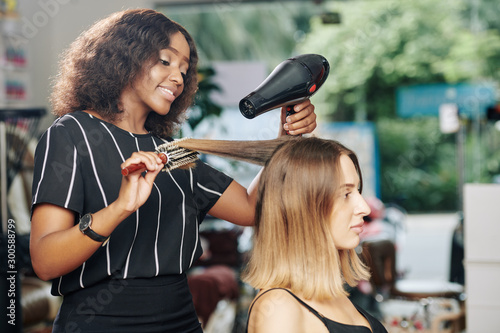 Positive young Black hairdresser enjoying working in beauty salon and blowdrying hair of client