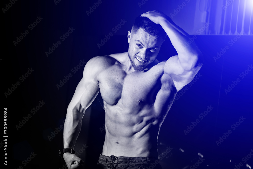 Naklejka premium Handsome man with big muscles, posing at the camera in the gym, Black and blue background. Portrait of a bodybuilder with naked torso. Closeup.