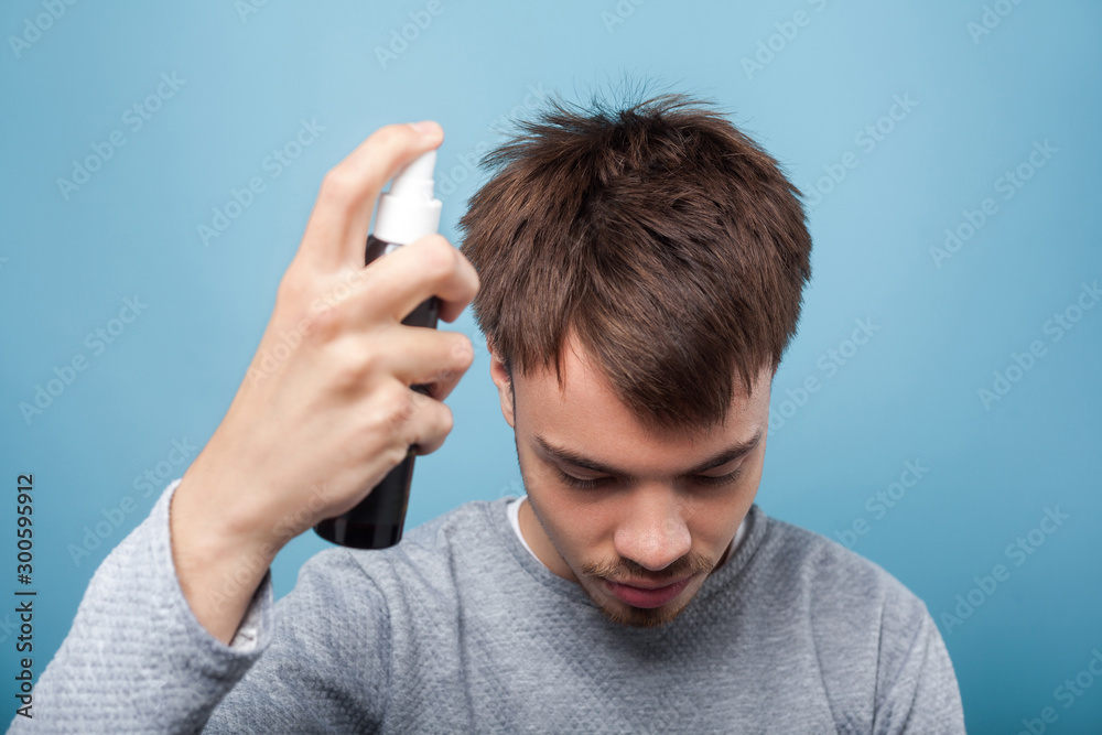 Portrait of young man in casual wear with head down, applying spray to his hair, treating premature hair loss or dandruff, healthcare and treatment. indoor studio shot isolated on blue background