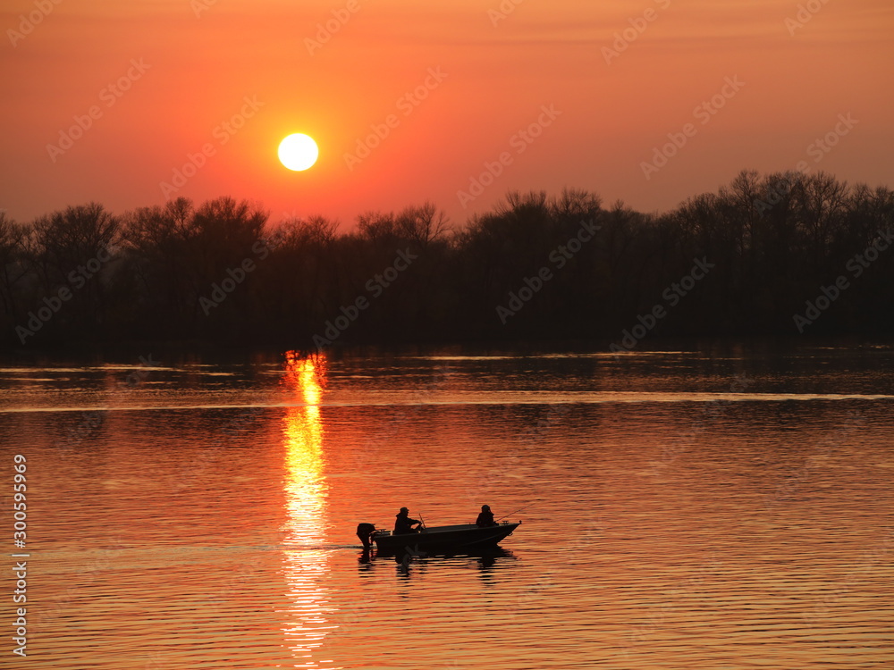 Φωτογραφία Two fishermen, a man and a woman, swim in a motor boat on the river against the background of an orange sunset