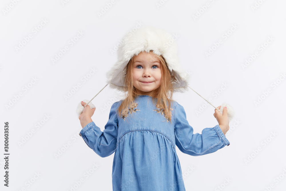 Smiling little surprised girl in a blue dress and a winter white fur hat having fun on White isolated background Smiling little surprised girl in a blue dress and a winter white fur hat having fun on White isolated background