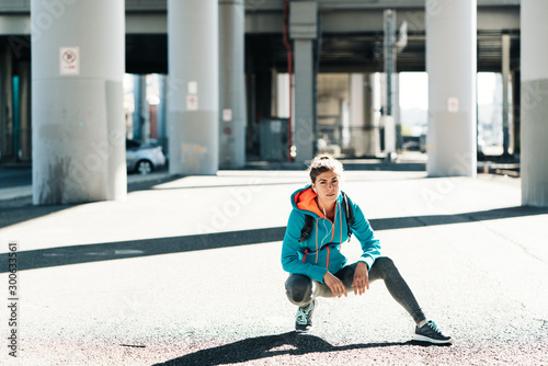 Portrait of young sporty woman sitting and resting under the bridge after workout in industrial city