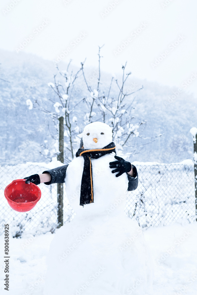   Winter teenager in a jacket and hat sculpts a snowman on a winter background with falling snow in frozen day concept
