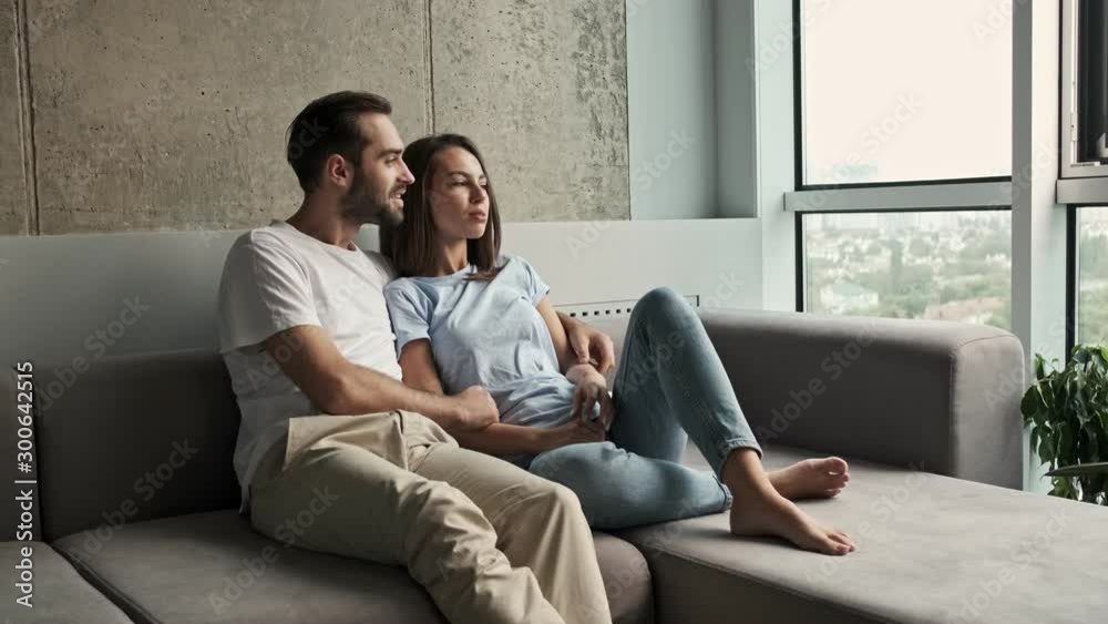 Beautiful young lovely couple hugging and talking while looking at the window sitting on sofa at home Beautiful young lovely couple hugging and talking while looking at the window sitting on sofa at home