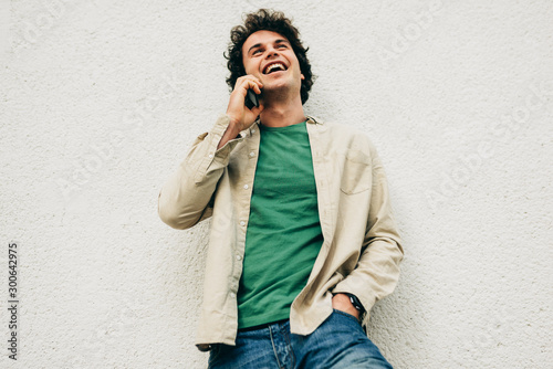 Outdoor portrait of young man talking on mobile phone with his friend. Happy male with curly hair resting outside making a call on his cell phone in the city street. Lifestyle, people