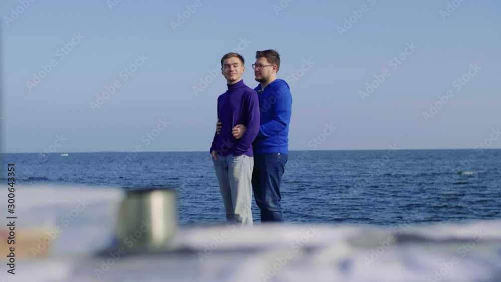 a picnic on the sea two men from the LGBT community, one approaches his boyfriend, hugs him, they look at each other and smile, shifting focus from the litter on which butyrbrod and a thermos a picnic on the sea two men from the LGBT community, one approaches his boyfriend, hugs him, they look at each other and smile, shifting focus from the litter on which butyrbrod and a thermos