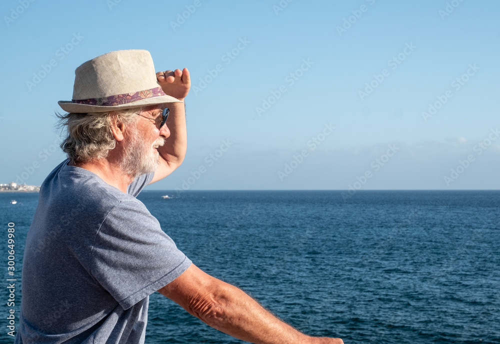 A senior man with white beard and hair looking far away at the sea from the cliff. Horizon over water. Blue sky and ocean on background