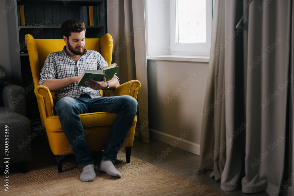 Confident young adult man with a beard reading a book by the window. Concept of everyday life.
