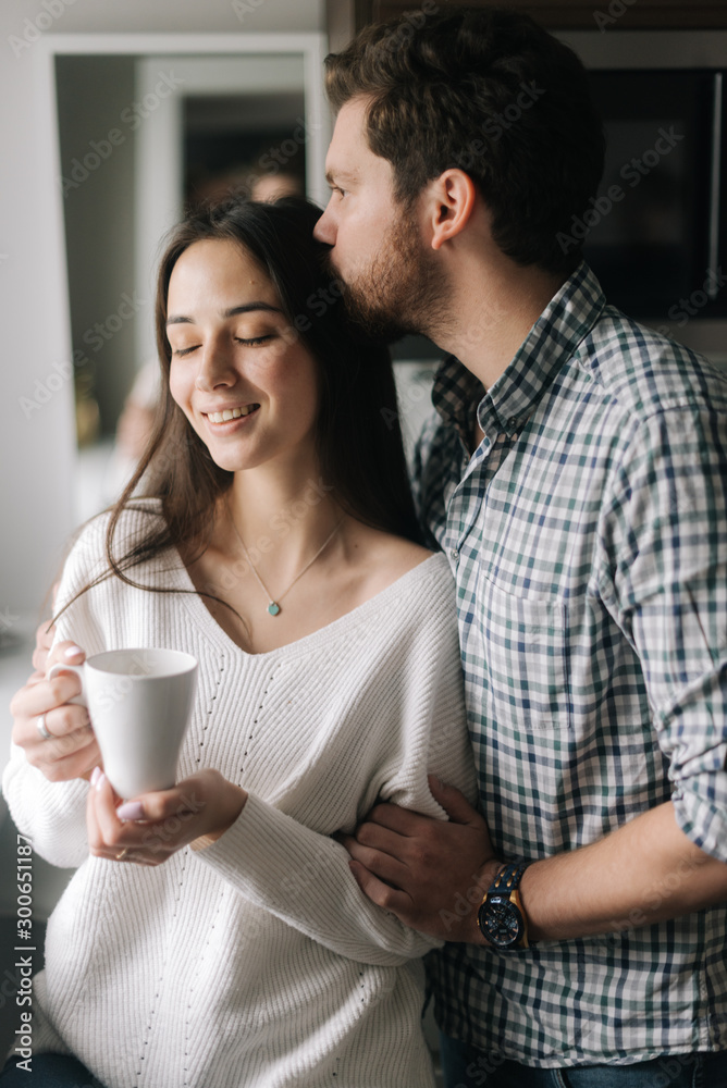 Handsome guy is kissing his girlfriend in th kitchen by the window. Daily life of a male and female in apartment. Handsome guy is kissing his girlfriend in th kitchen by the window. Daily life of a male and female in apartment.