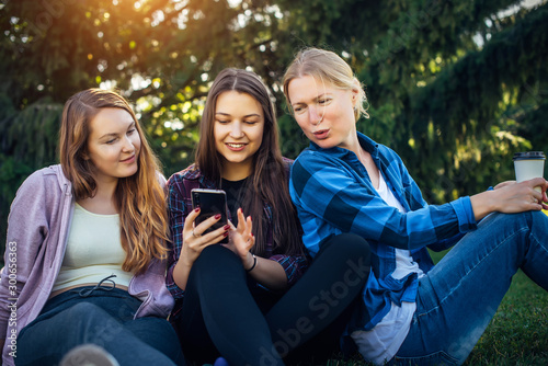 Three cute girls relax and socialize on the lawn in summer park. Young women sit on the green grass among the trees and look into the smartphone. Students in between classes outdoor, close up.