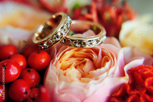 Gorgeous wedding rings with patterns on a bright wedding bouquet  close-up. Two gold rings on a blurred background of scarlet flowers.