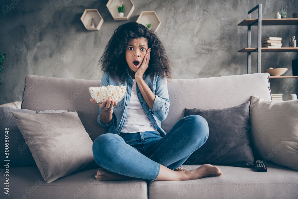 Photo of pretty dark skin wavy lady homey mood eating popcorn watching television horror show hand on cheek sitting cozy couch casual jeans outfit flat indoors - obrazy, fototapety, plakaty 