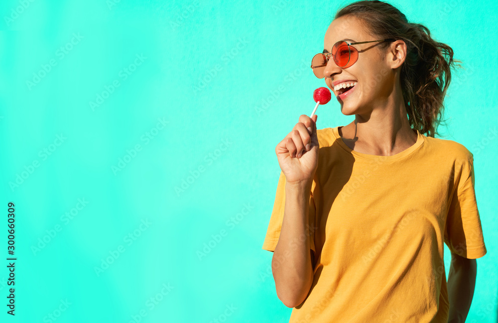 Φωτογραφία portrait of a young stylish laughing emotional woman with big red lollipop, standing against blue background