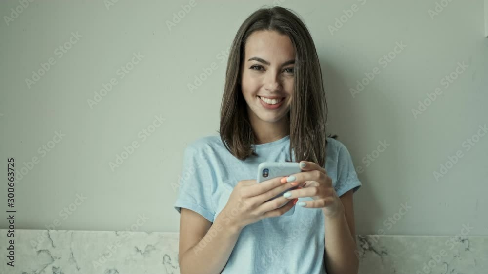 Cute cheerful young brunette woman smiling and laughing while chatting on smartphone standing near the wall at home Cute cheerful young brunette woman smiling and laughing while chatting on smartphone standing near the wall at home