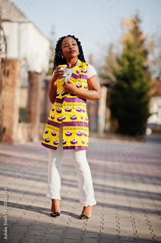 Stylish african american women in yellow jacket posed on street with hot drink in disposable paper cup.