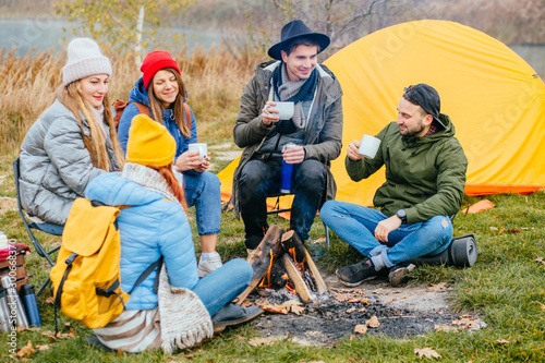 Group of five people traveler wearing warm colorful clothes sitting around bonfire in camp. Best friends warms and drinking hot tea  joking  laughing together in autmn time nature outdoor.