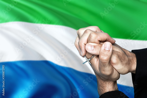 Sierra Leone flag and praying patriot man with crossed hands. Holding cross, hoping and wishing. © sezerozger