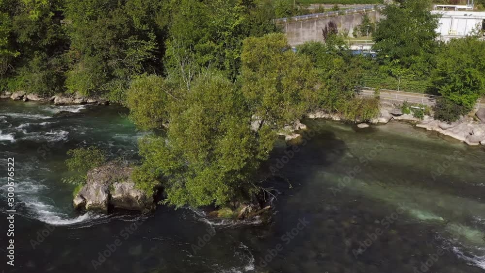 Tracking aerial shot of a tree sitting on a rock in the river Rhine filled with green boisterous mountain water. Tracking aerial shot of a tree sitting on a rock in the river Rhine filled with green boisterous mountain water.