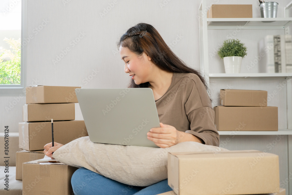 Young entrepreneur, sitting on the sofa and working at home. Checking orders from customers by laptop and writing customer address deliver shipment online sales. Concept of small business owner. Young entrepreneur, sitting on the sofa and working at home. Checking orders from customers by laptop and writing customer address deliver shipment online sales. Concept of small business owner.