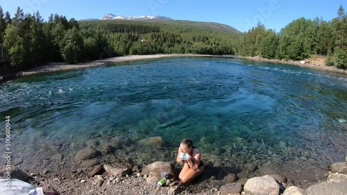 A young, attractive, brunette woman in a swimsuit is washing herself with a loofah and soap in a blue and turquoise colored river in the wild outdoors in Norway.