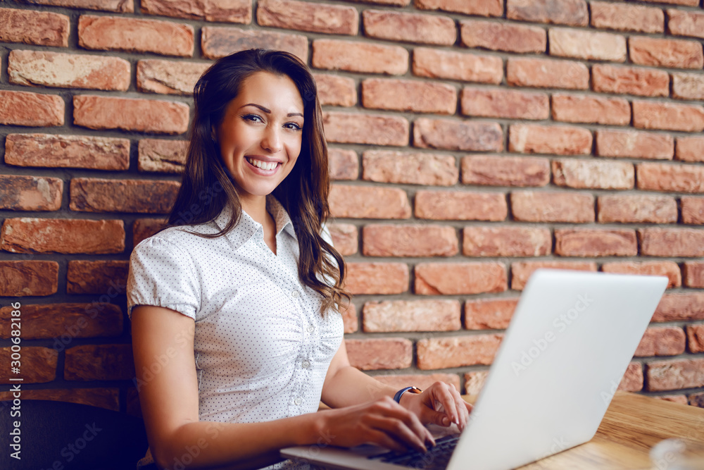 Happy pretty caucasian brunette sitting in cafe and using laptop. Hands are on keyboard. In background is brick wall. Happy pretty caucasian brunette sitting in cafe and using laptop. Hands are on keyboard. In background is brick wall.