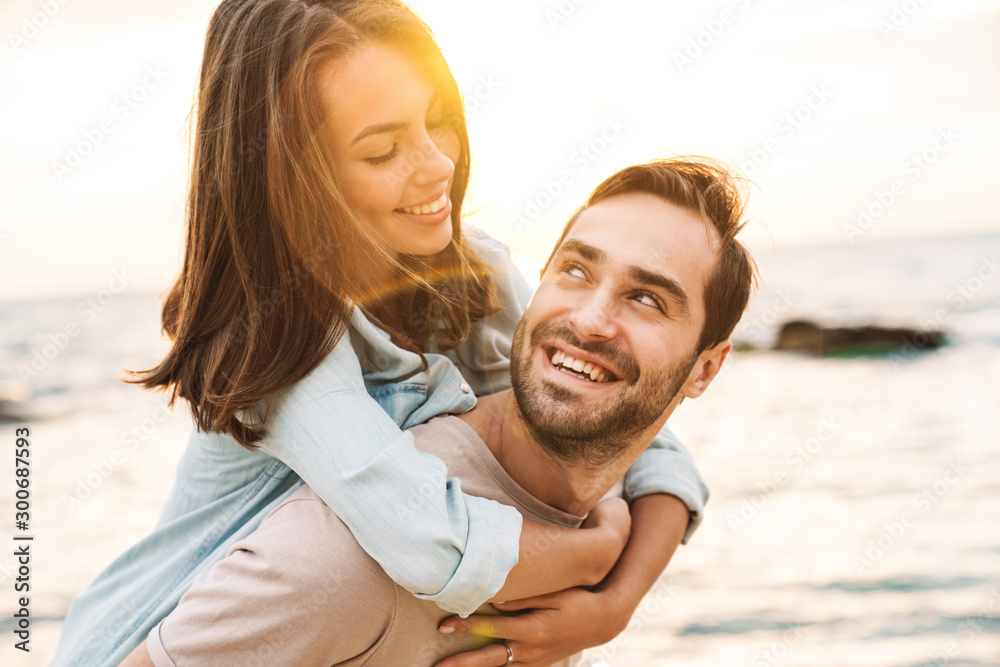Image of young man giving piggyback ride and looking at beautiful woman - obrazy, fototapety, plakaty 