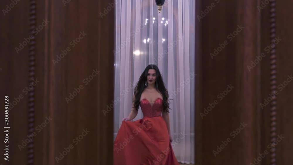 A girl in a red dress in a photo studio looks like a 19th-century Russian palace with high ceilings, she opens the door and goes to the window, moving slowly. She has a smile on her face. A girl in a red dress in a photo studio looks like a 19th-century Russian palace with high ceilings, she opens the door and goes to the window, moving slowly. She has a smile on her face.