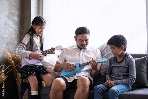 Father sitting at the middle of daughter and son,playing ukulele together,relax time,with happy feeling,at living room,blurry light around